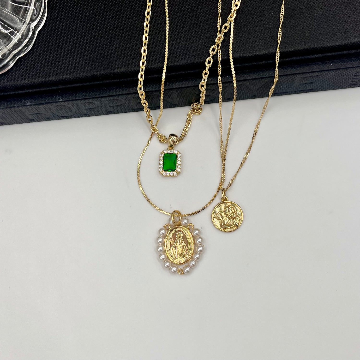 3 necklaces laid out on a white background. Emerald pendant on a gold chain. Saint Mary with pearls surrounding it on a gold chain. Baby angel pendant on a gold chain